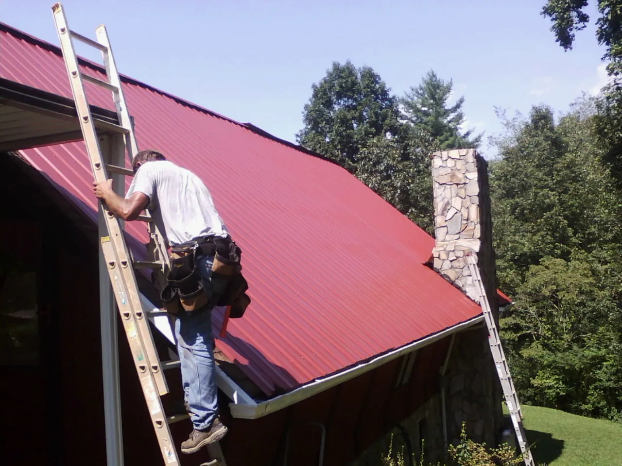 Roofer on a ladder beside a red metal roof and stone chimney — Soffit & Fascia Repair by Roofing Brothers in Sedgwick